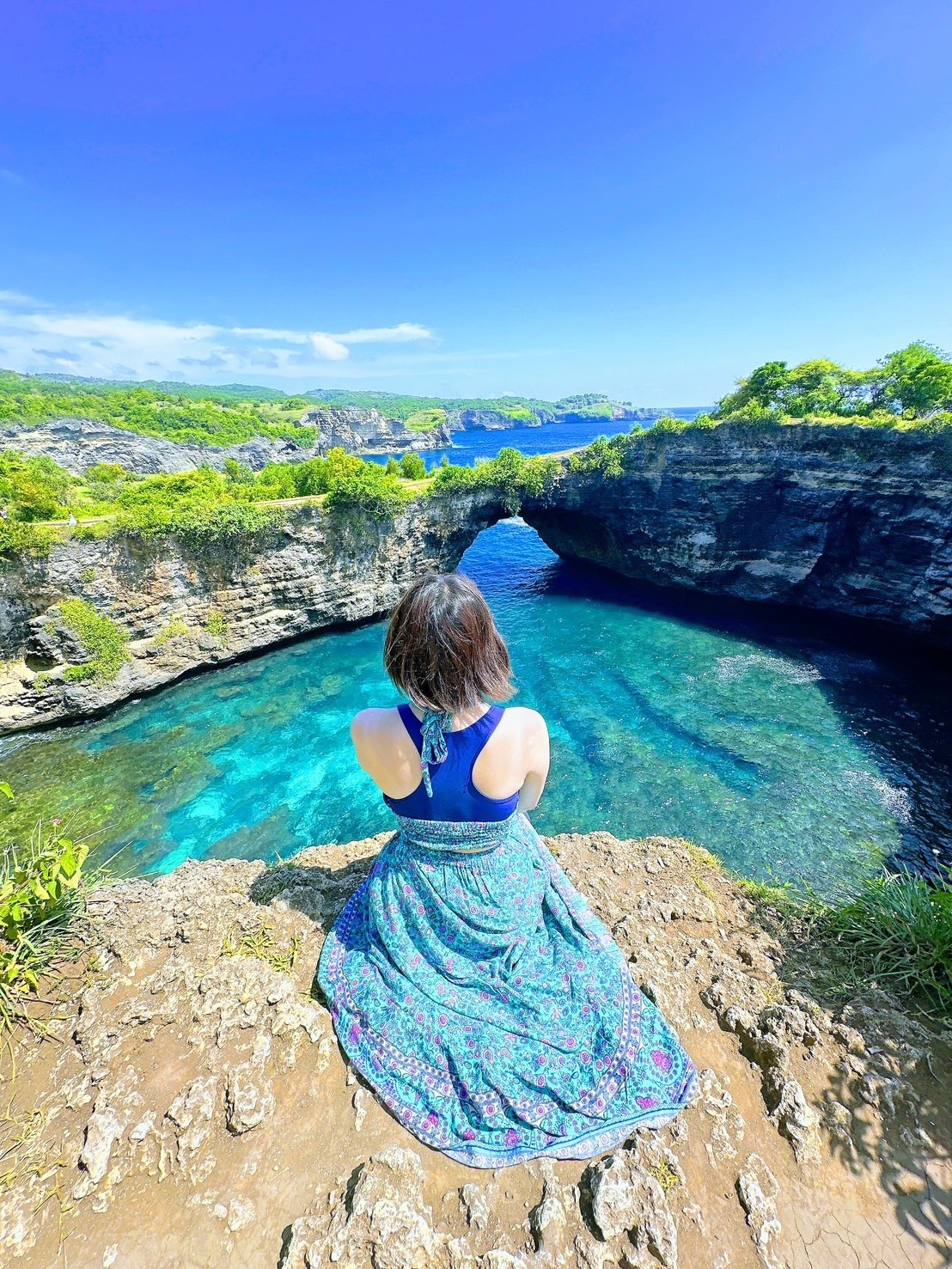 岩場から青い海と天然の橋を眺めるCHIAKIの後ろ姿